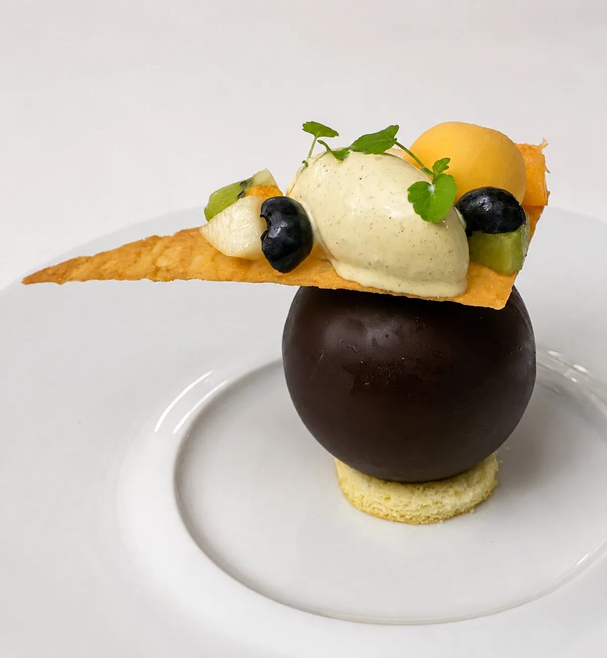 Un dessert original composé d’une sphère en chocolat, d’une tuile croustillante, de fruits et de glace sur une assiette blanche