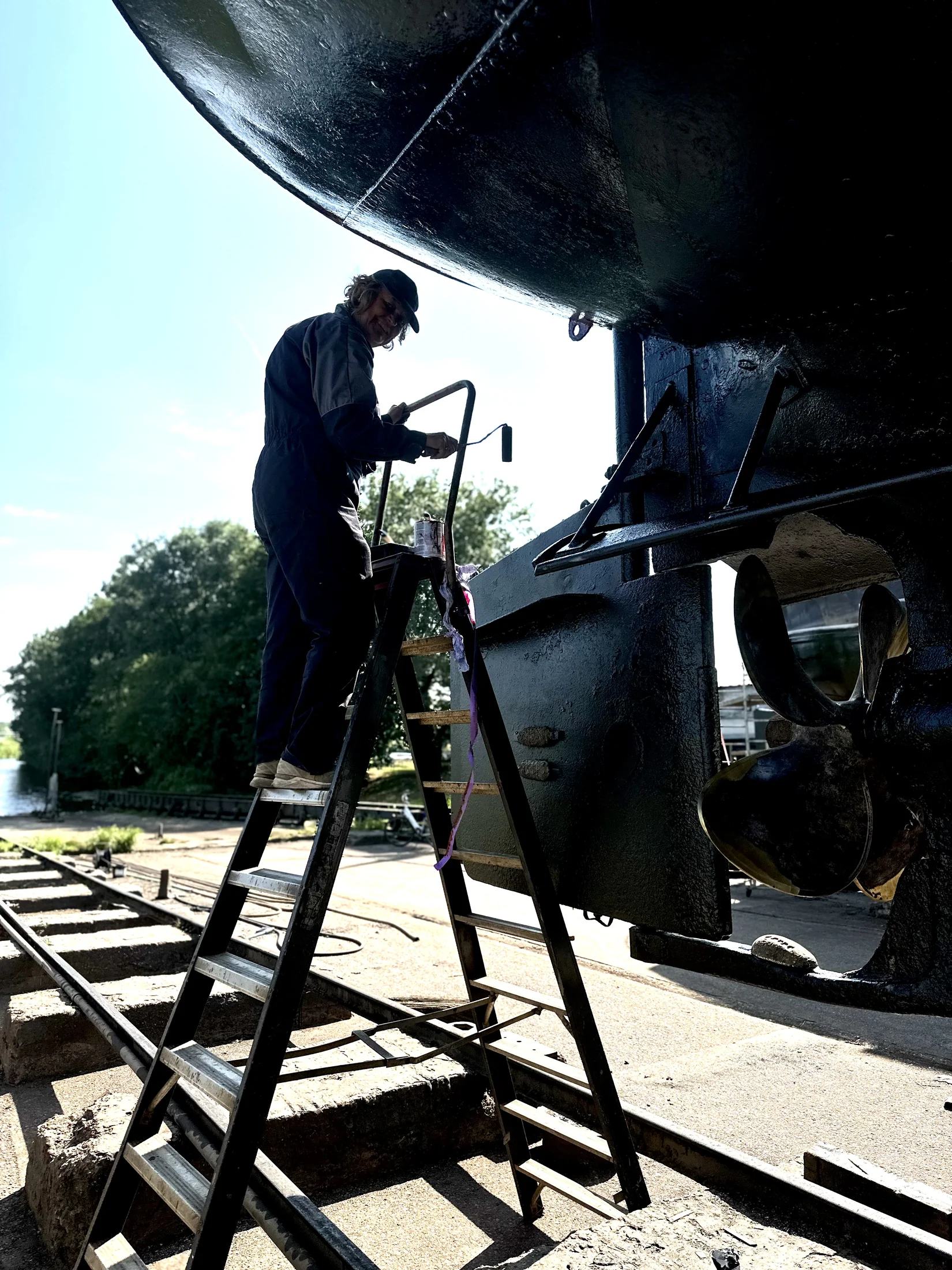 Vanaf een ladder wordt op de werf in Coevorden een zwarte coating onder Maria’s achtersteven aangebracht