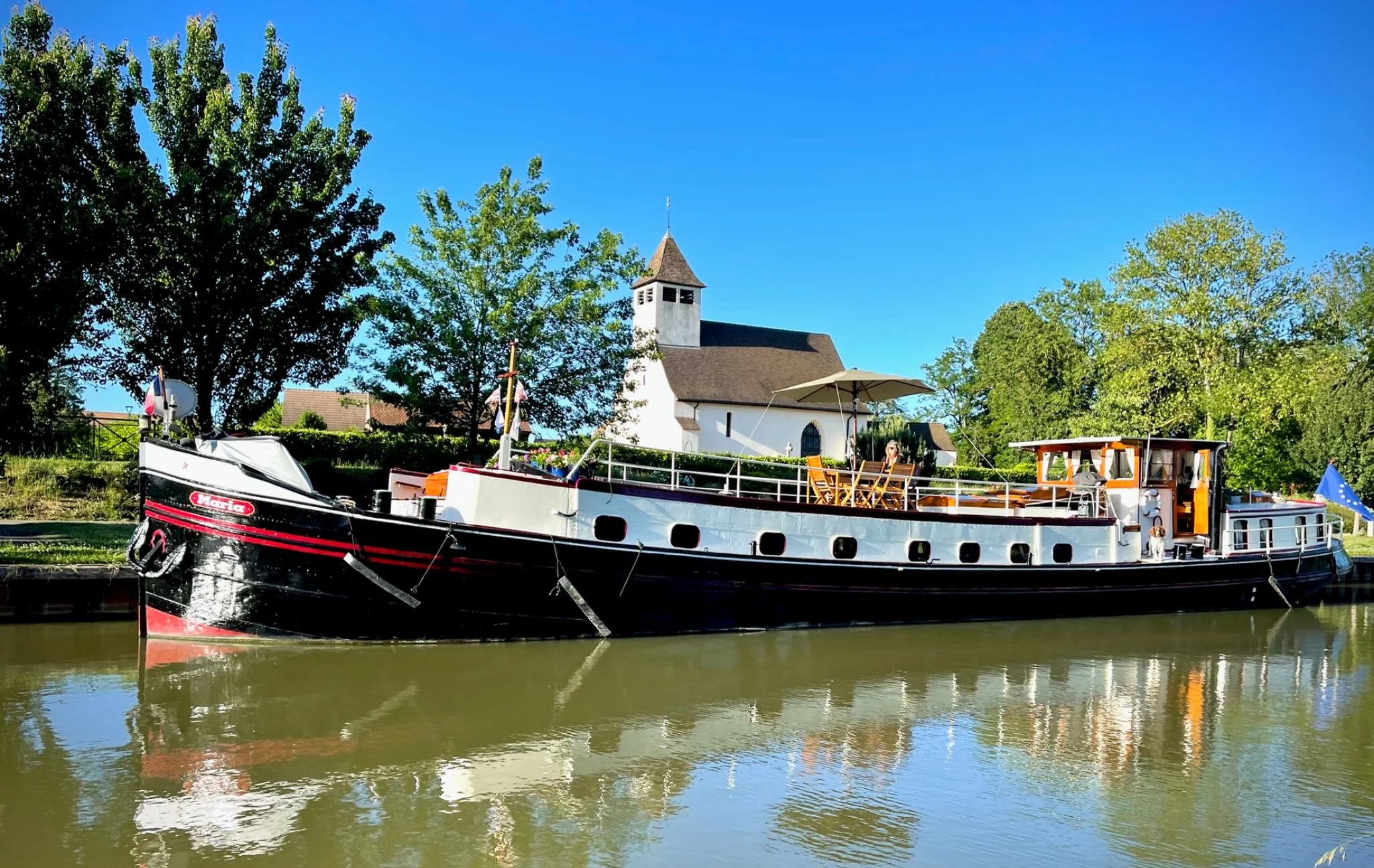 Maria vue de profil dans son ensemble, près d’une petite église au bord de l’eau